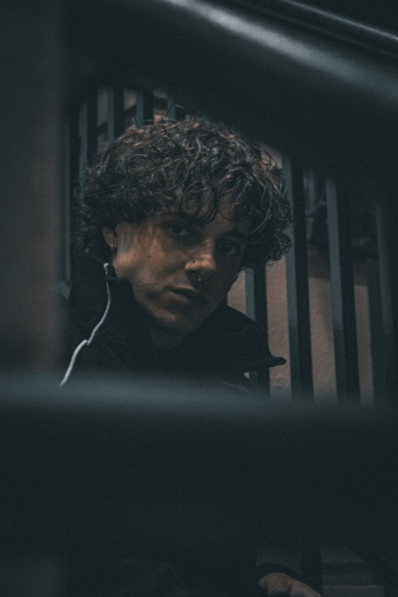 Person with curly hair and a nose ring sitting on stairs in dim light.