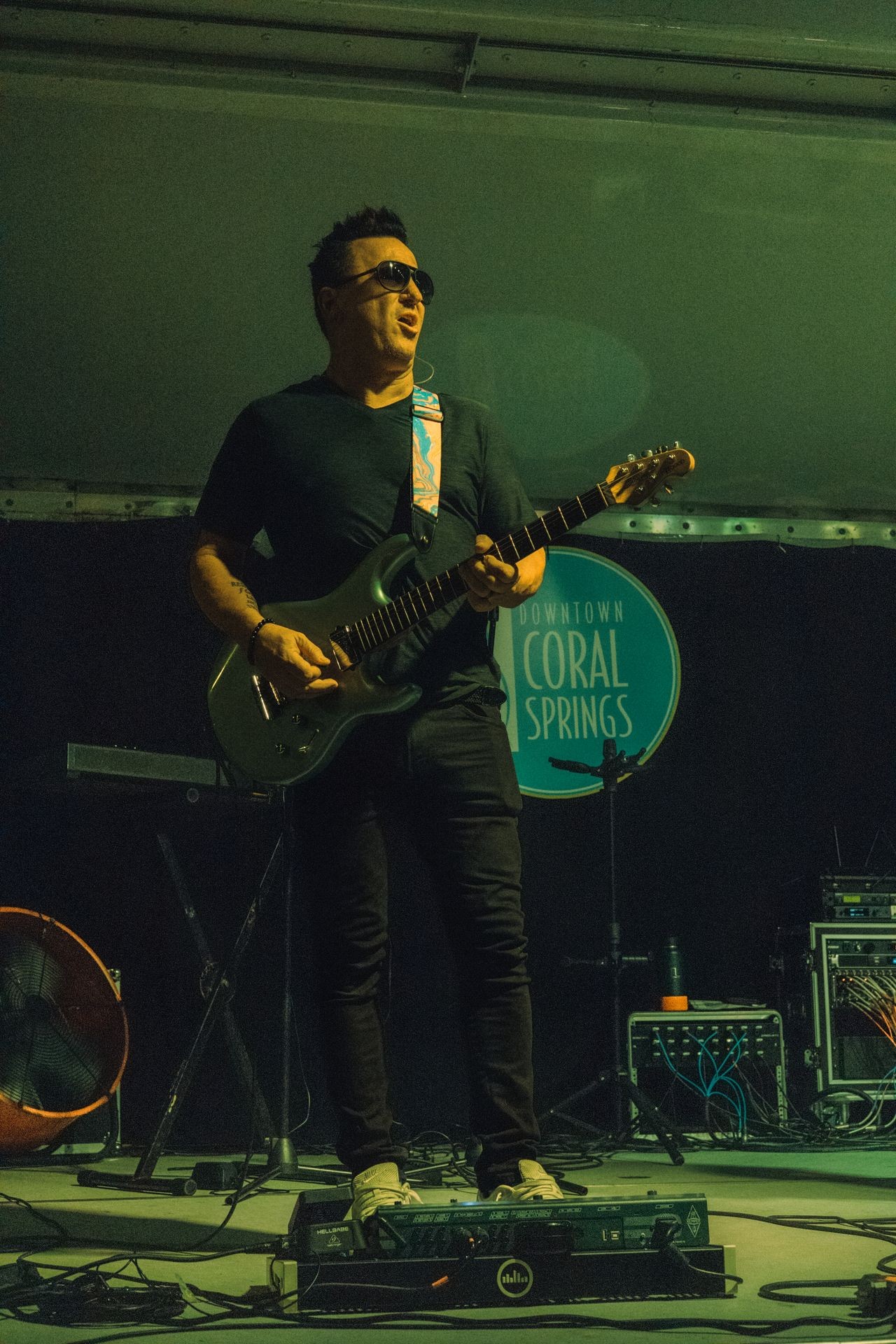 Musician performing on stage with electric guitar and pedalboard, under green lighting.